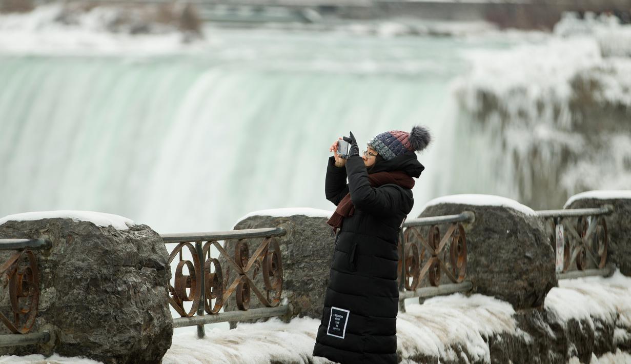 Seorang turis berjaket tebal mengambil gambar di depan Air Terjun Niagara, Ontario, Kanada, Senin (9/1). Salju tebal dan angin dingin membahayakan kondisi warga. (Geoff Robins/AFP)