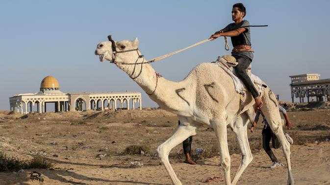 Seorang joki Palestina beraksi saat bersaing dalam perlombaan unta lokal yang diadakan di lokasi bekas bandara Gaza yang hancur, di Rafah, Jalur Gaza selatan (20/10/2019). Bandara Internasional Yasser Arafat yang diresmikan November 1998, kini tinggal reruntuhan akibat serangan Israel. (AFP Photo/Sa