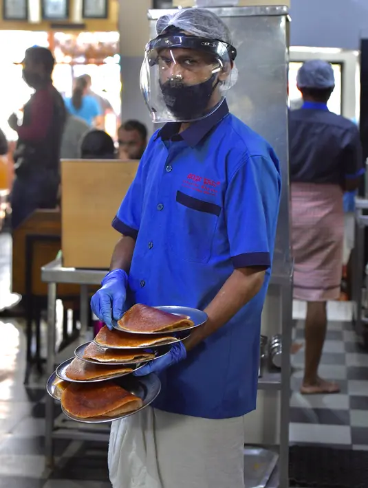 Pramusaji mengenakan pelindung wajah dan masker di sebuah restoran yang dibuka kembali usai pelonggaran lockdown di Bangalore, India, Senin (8/6/2020). Untuk keamanan pelanggan, restoran-restoran menyiapkan panel transparan di meja makan sebagai pencegahan COVID-19. (Manjunath Kiran/AFP)