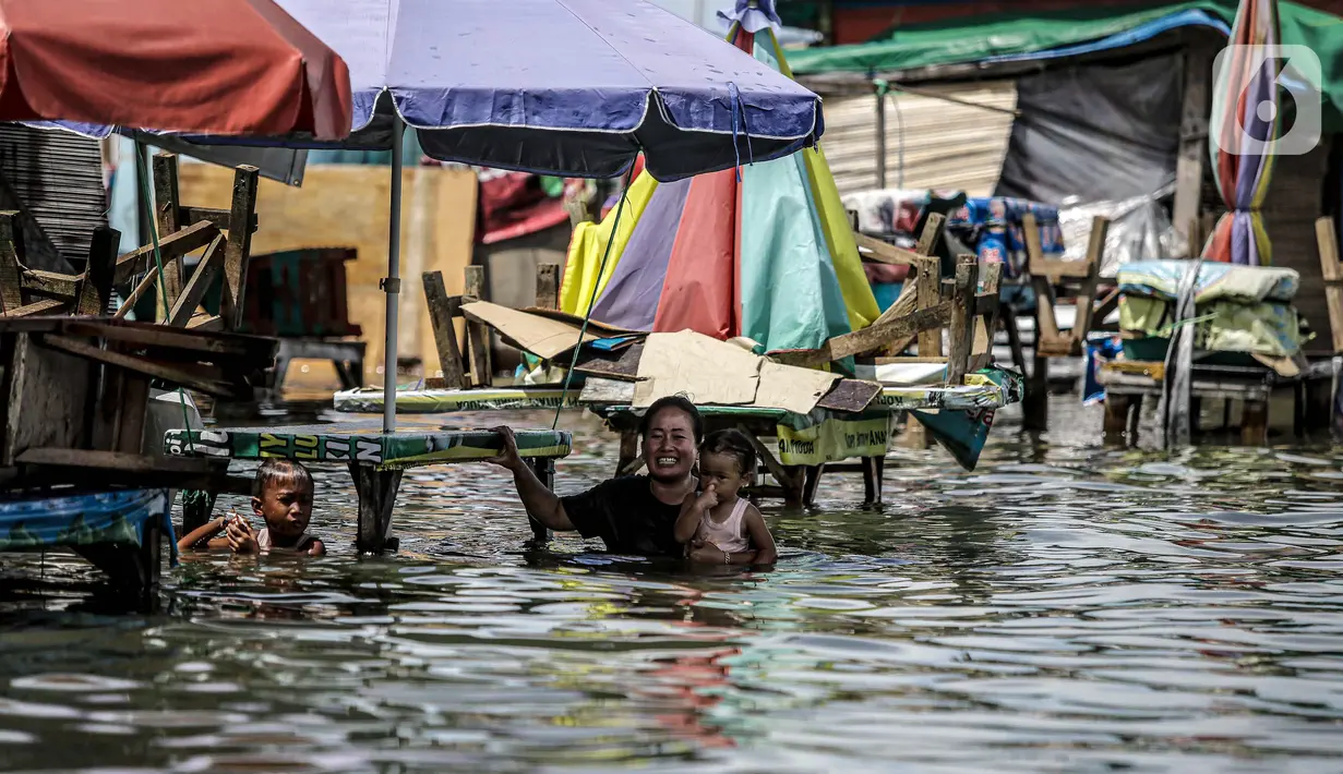 Seorang wanita menggendong anak kecil melintasi banjir rob di kawasan Muara Angke, Jakarta, Selasa (9/11/2021). Badan Penanggulangan Bencana Daerah (BPBD) DKI Jakarta memperkirakan puncak musim hujan dan potensi rob di Ibu Kota terjadi pada Januari hingga Februari 2022. (Liputan6.com/Faizal Fanani)