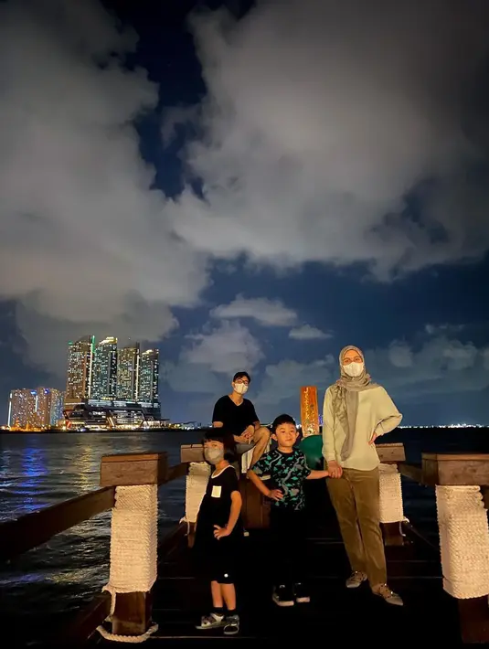 Pasangan Nycta Gina dan Rizky Kinos beserta kedua anaknya foto di pantai dengan latar belakang gedung bertingkat. (Instagram/missnyctagina)