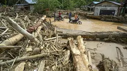 Tumpukan material kayu yang terbawa air saat banjir bandang tidak hanya menutup jalan, tetapi juga menimbun rumah dan halaman warga. Tampak dalam foto, warga desa mengarungi banjir di jalan yang tergenang akibat banjir bandang di Desa Tukka, Tapanuli Tengah, Sumatera Utara, pada 2 Desember 2025. (YT HARIONO/AFP)