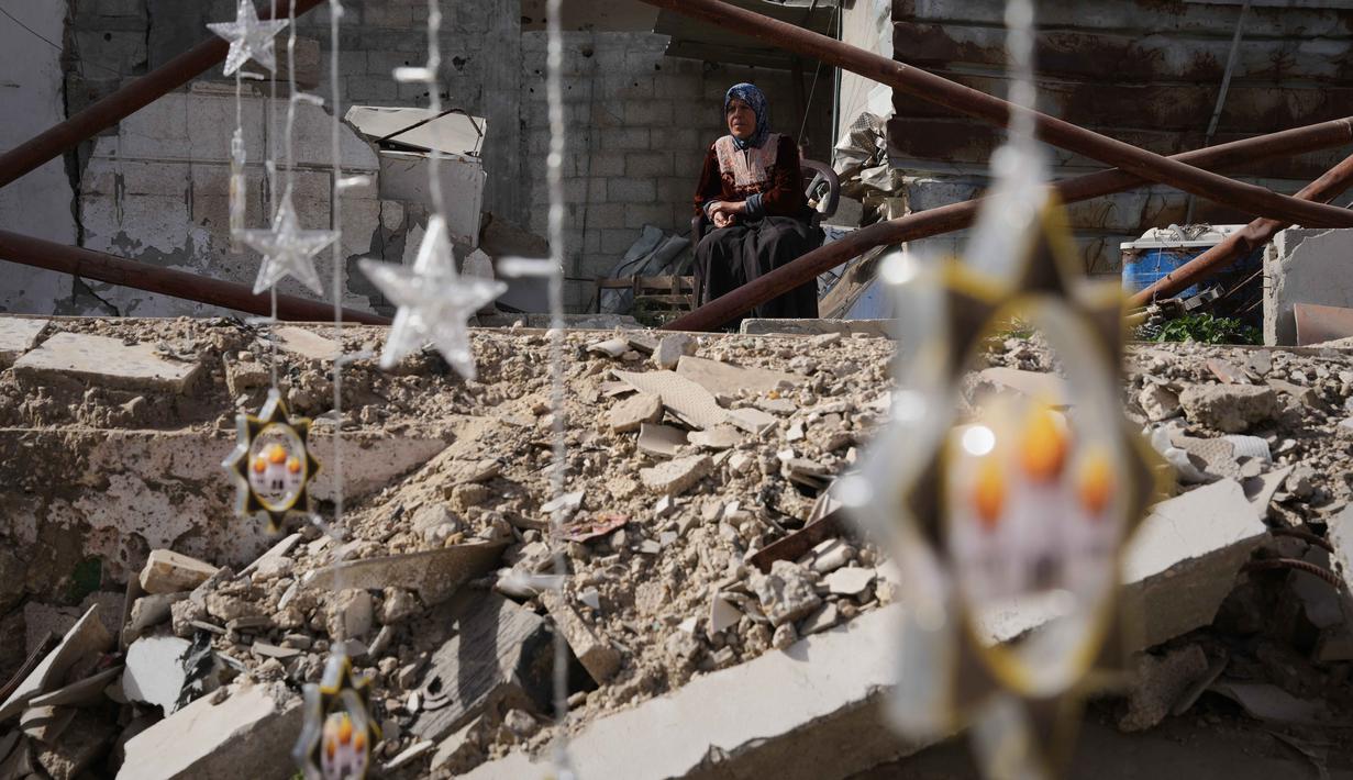 Beberapa pengungsi membuat lentera Ramadan dari kaleng bekas atau kain selimut bantuan. Tampak dalam foto, seorang wanita Palestina memperhatikan orang lain menggantung hiasan Ramadan di samping reruntuhan rumah yang hancur di Khan Younis, Jalur Gaza selatan, Minggu 15 Februari 2026. (AP Photo/Abdel Kareem Hana)