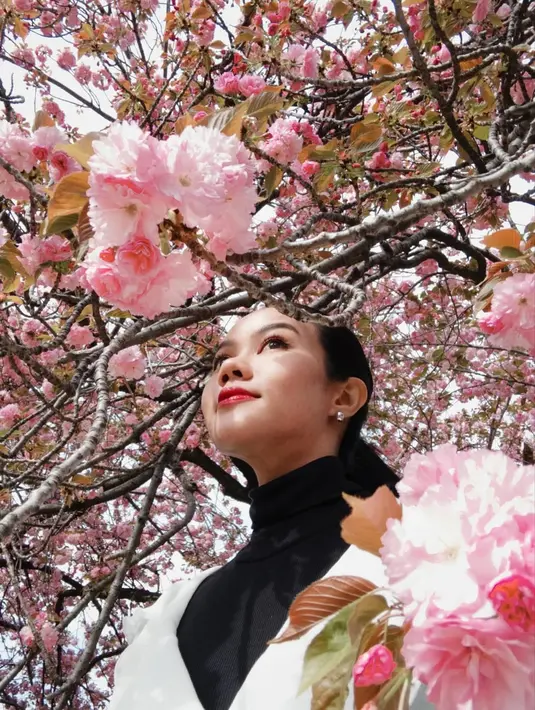 Rambut panjangnya dibiarkan diikat ke belakang, tertiup angin, menciptakan kesan natural namun tetap anggun. Riasan wajahnya pun tampak minimalis namun tetap memikat dengan pulasan lipstik merah yang menjadi statement tersendiri, membuat penampilannya semakin stand out di antara hamparan bunga sakura berwarna pink pastel. [@yurayunita].