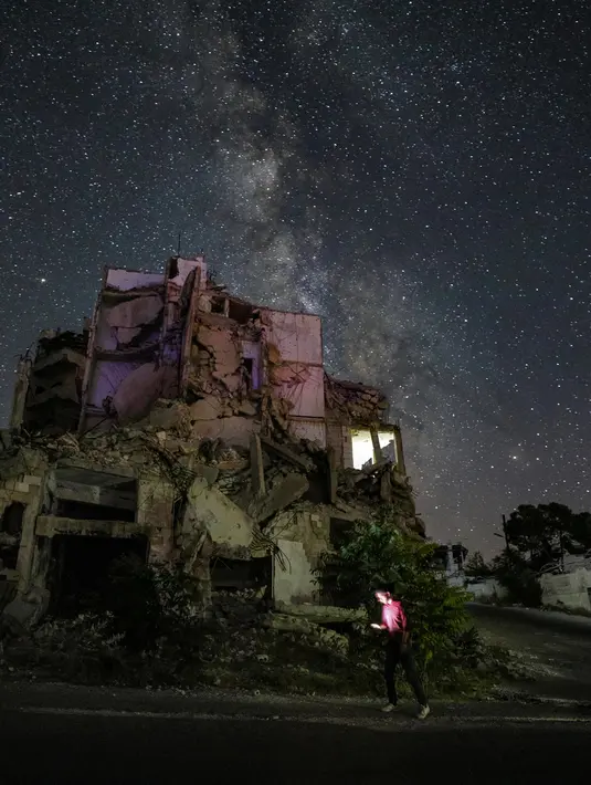 Galaksi Bima Sakti menghiasi langit malam saat seorang pria berjalan melewati gedung-gedung yang hancur akibat pengeboman di Kota Ariha, Provinsi Idlib, Suriah, Sabtu (27/6/2020). (Omar HAJ KADOUR/AFP)