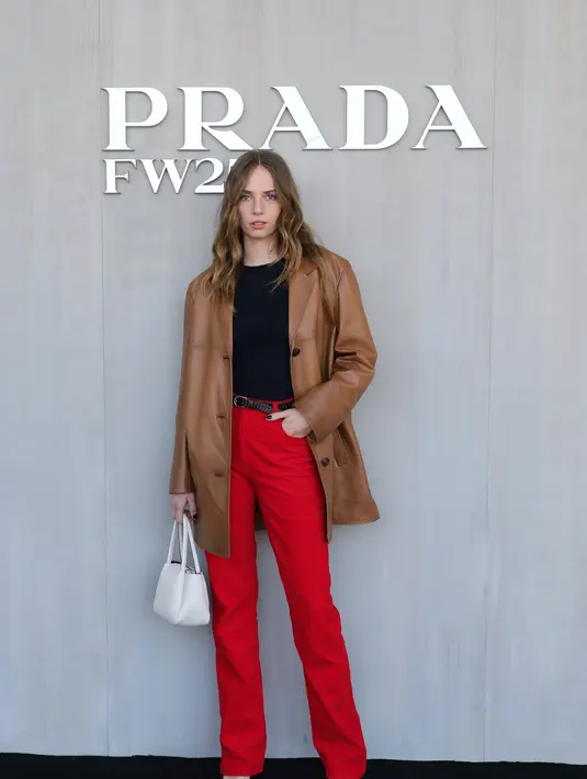 Maya Hawke memberikan sentuhan bold pada tampilan semi formalnya. Ia memadukan leather blazer coklat dengan high waisted pants merah dan kaus hitam. [Dok/Prada]