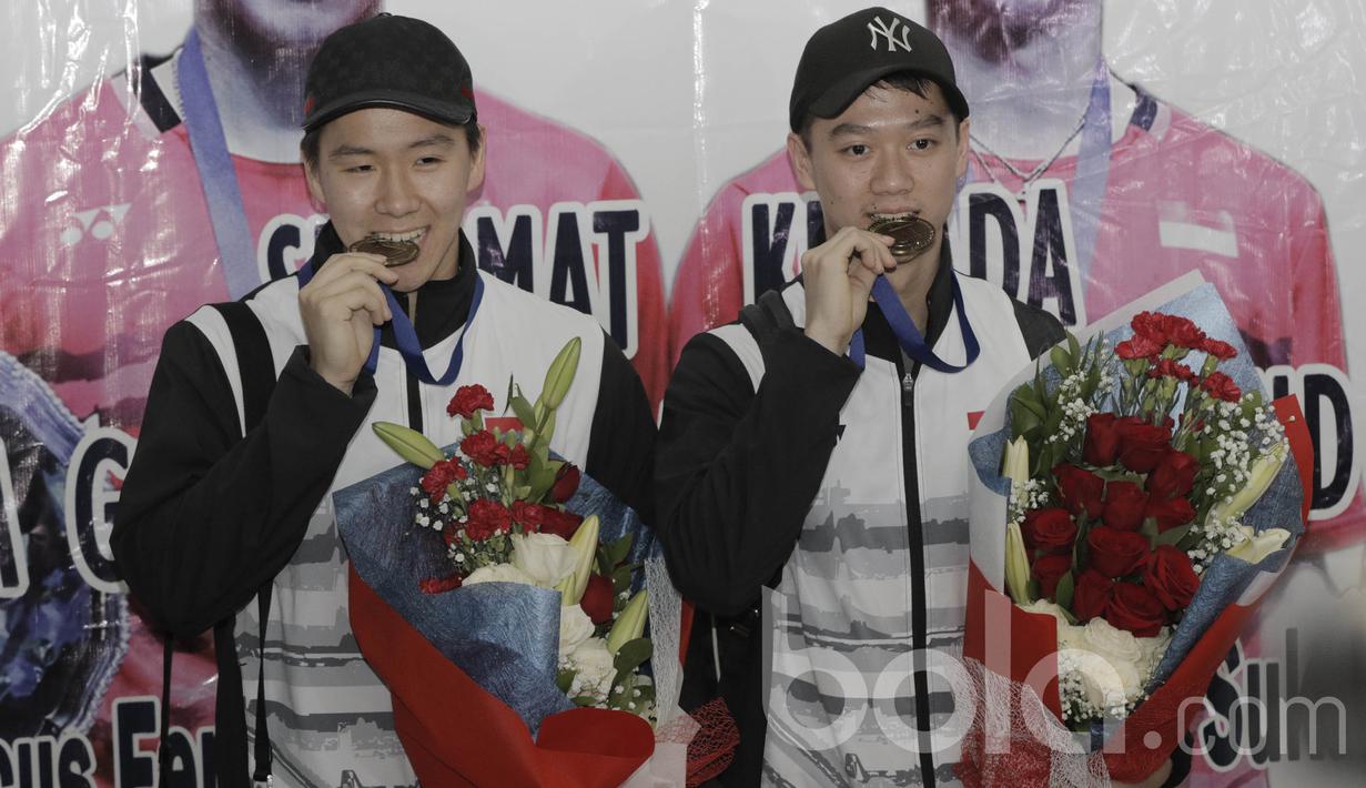 Pebulutangkis ganda putra Indonesia, Kevin Sanjaya/Marcus Gideon, menggigit medali saat tiba di Bandara Soekarno Hatta, Banten, Selasa (14/3/2017). Pasangan ini kembali setelah berhasil meraih gelar All England. (Bola.com/M Iqbal Ichsan)