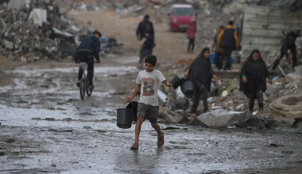 Diketahui, sekitar 85% infrastruktur air dan sanitasi di Gaza telah rusak atau hancur akibat serangan militer Israel dalam dua tahun terakhir. Tampak dalam foto, seorang anak laki-laki membawa dua ember kosong saat berjalan menuju tempat mengambil air di tengah hujan deras di kawasan Sheikh Radwan, Kota Gaza, Jumat 14 November 2025. (AP Photo/Jehad Alshrafi)