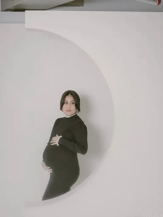Ia tampil cantik berbalut bodycon dress warna hitam dengan aksen putih pada tepi dress. Busana yang dikenakan bumil satu ini sangat kontras dengan latar studio yang serba putih. (Instagram/gyaps).