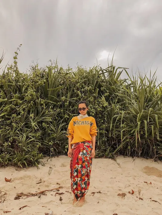 Indah berpose di sebuah pantai menggunakan sweater kuning dan batik berwarna merah bercorak. Ditambah kacamata hitam, dia tampak percaya diri dengan background semak dan langit kelabu. (Liputan6.com/IG/indahpermatas)