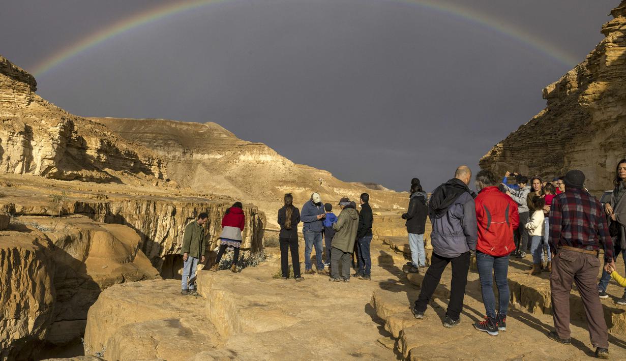 Pelangi terlihat saat warga Israel melihat banjir bandang yang terjadi menyusul hujan lebat di gurun Negev di sebelah timur kota Dimona Israel pada 26 Desember 2022. Hujan deras mengguyur Israel utara, dataran pantai, Yerusalem, dan sebagian Negev utara, termasuk kilat dan angin kencang di beberapa daerah. (AFP/Menahem Kahana)