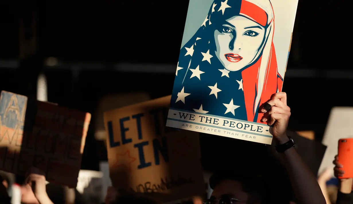 Pengunjuk rasa membawa poster wanita berhijab bendera Amerika saat aksi protes di Bandara Internasional San Francisco, Sabtu (28/1). Mereka memprotes kebijakan Donald Trump yang melarang warga dari 7 negara muslim masuk AS. (AP Photo/Marcio Jose Sanchez)