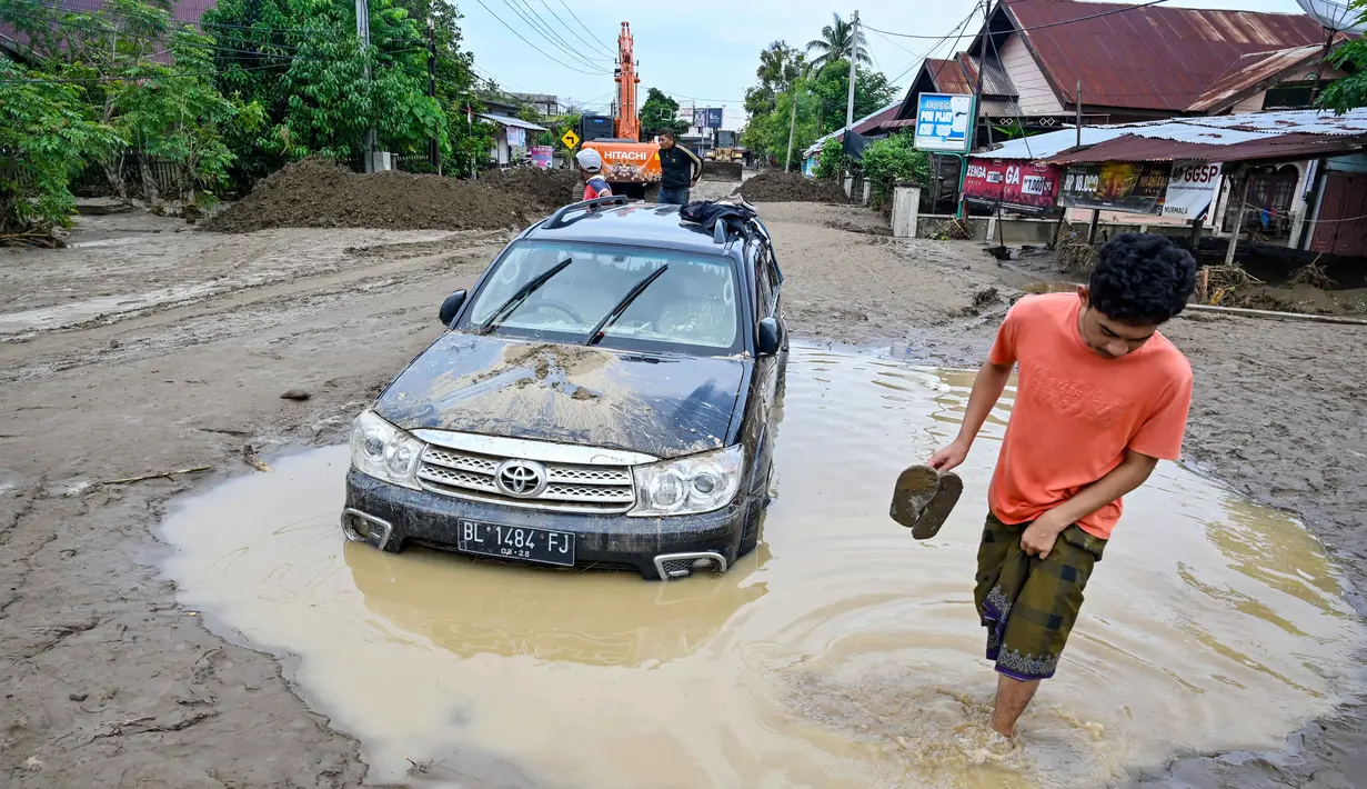 Sejumlah kendaraan roda dua dan roda empat milik warga juga ikut hanyut terbawa air sungai. Tampak dalam foto, warga berjalan melewati mobil yang terdampar di lumpur tebal akibat banjir bandang di Meureudu, Kabupaten Pidie Jaya, Provinsi Aceh, Jumat 28 November 2025. (CHAIDEER MAHYUDDIN/AFP)