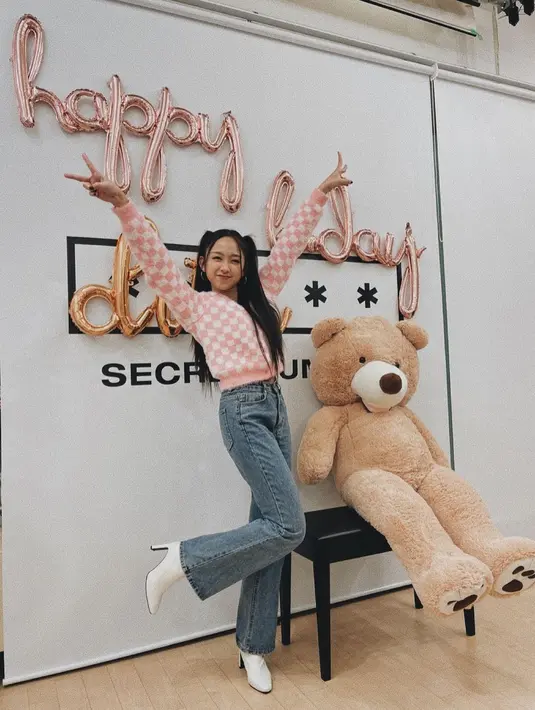 Berpose dengan boneka beruang super besar, Dita Karang tampil full makeup bernuansa oranye netral, mengikat dua rambutnya sedikit ke belakang, dan mengenakan outfit kasual. Wool sweater dengan motif kotak-kotak bernuansa merah muda dan putih dipadunya dengan celana jeans, serta boots heels putih.