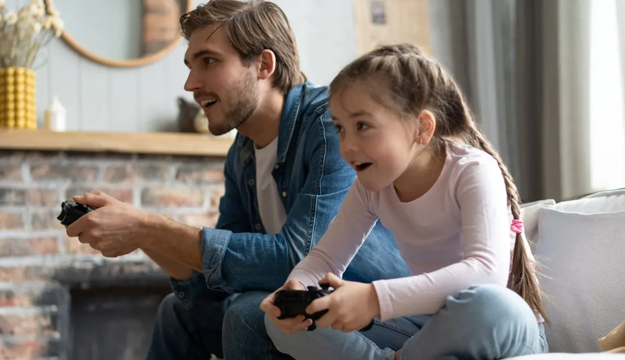 Seorang ayah bersama putrinya sedang bermain video game di dalam rumah, mereka terlihat begitu bersemangat mengalahkan satu sama lain. (OPOLJA/ Shutterstock.com)