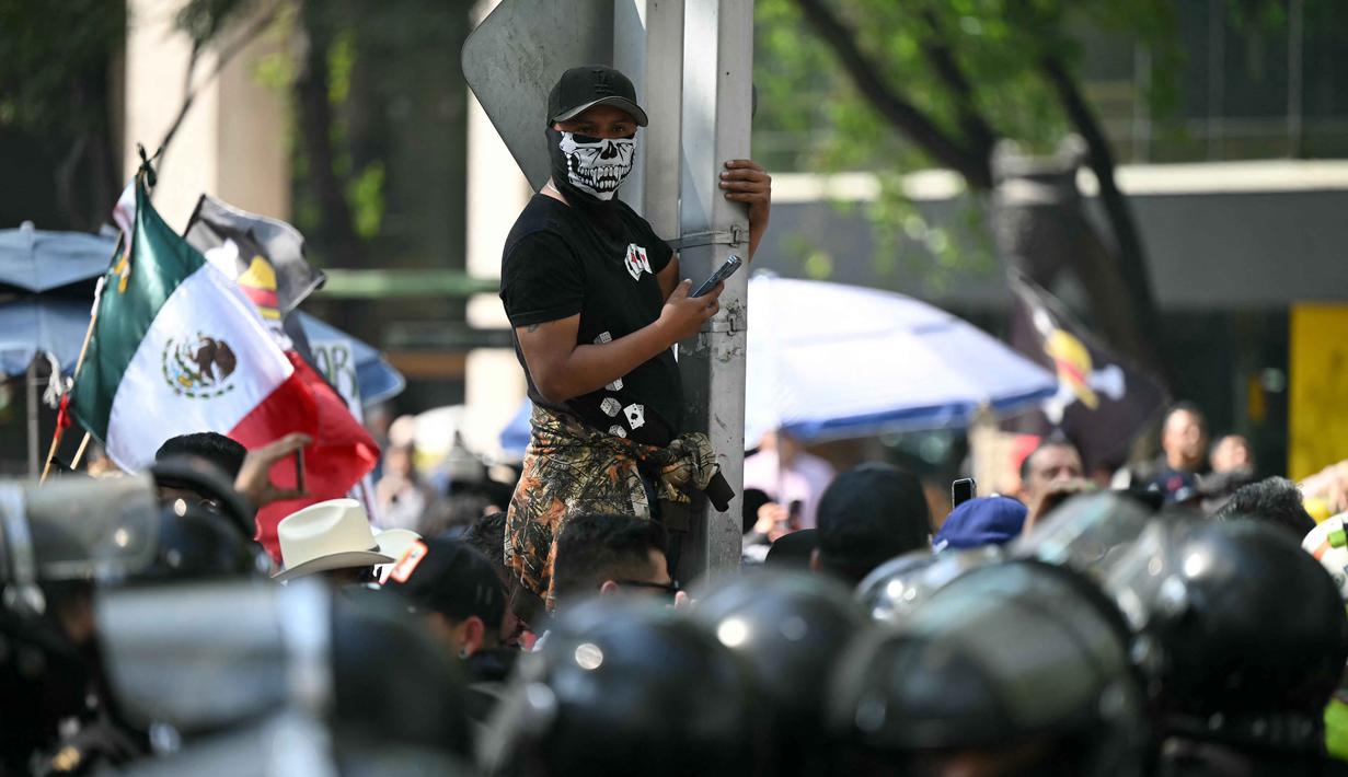 Pengunjuk rasa juga membawa simbol bendera bajak laut "One Piece". Tampak dalam foto, seorang pengunjuk rasa bertopeng tengkorak melihat ke arah polisi anti huru hara dalam demonstrasi Generasi Z melawan pemerintahan Presiden Meksiko Claudia Sheinbaum, di sepanjang Reforma Avenue di Mexico City, pada 20 November 2025. (Yuri CORTEZ/AFP)