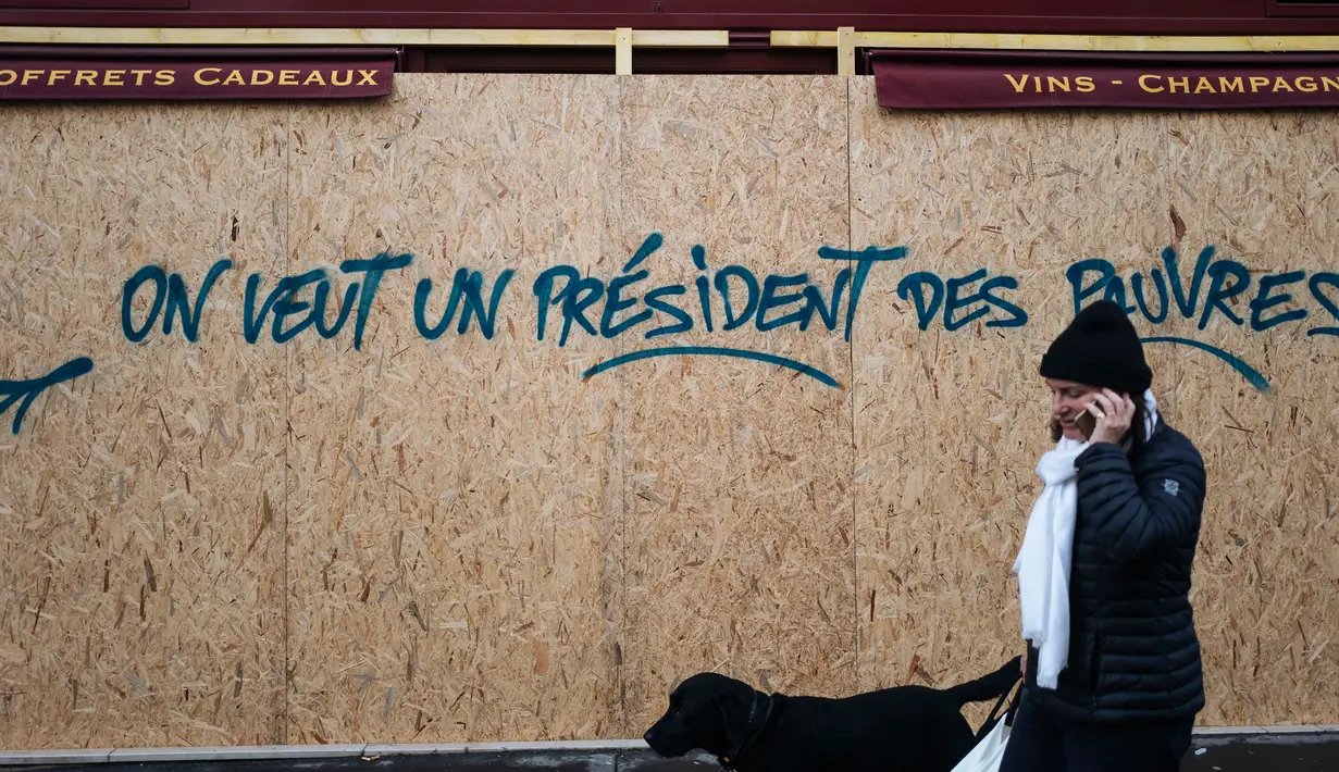 Seorang wanita berjalan melewati grafiti bertuliskan 'kami ingin seorang presiden untuk orang miskin' di sebuah toko dekat Arc de Triomphe, Paris, Prancis,  Minggu (2/12). Tiga orang tewas dalam unjuk rasa sejak dua pekan lalu. (AP Photo/Kamil Zihnioglu)