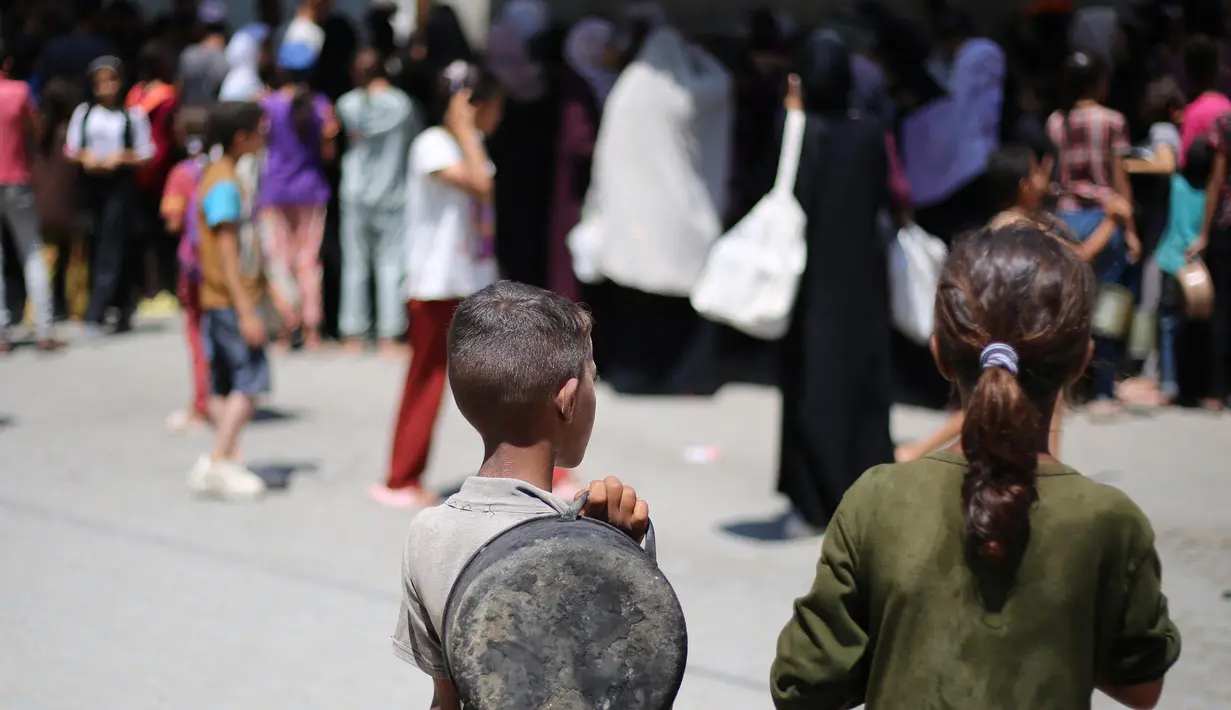 Anak-anak Palestina menyaksikan orang-orang mengantre untuk mendapatkan makanan hangat yang dibagikan oleh dapur amal di kamp pengungsi Nuseirat, Jalur Gaza tengah pada 15 Juli 2025. (Eyad BABA/AFP)