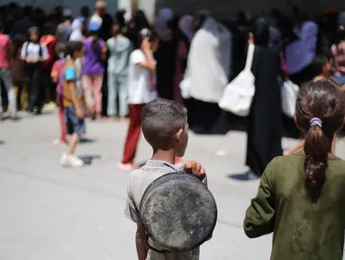 Anak-anak Palestina menyaksikan orang-orang mengantre untuk mendapatkan makanan hangat yang dibagikan oleh dapur amal di kamp pengungsi Nuseirat, Jalur Gaza tengah pada 15 Juli 2025. (Eyad BABA/AFP)
