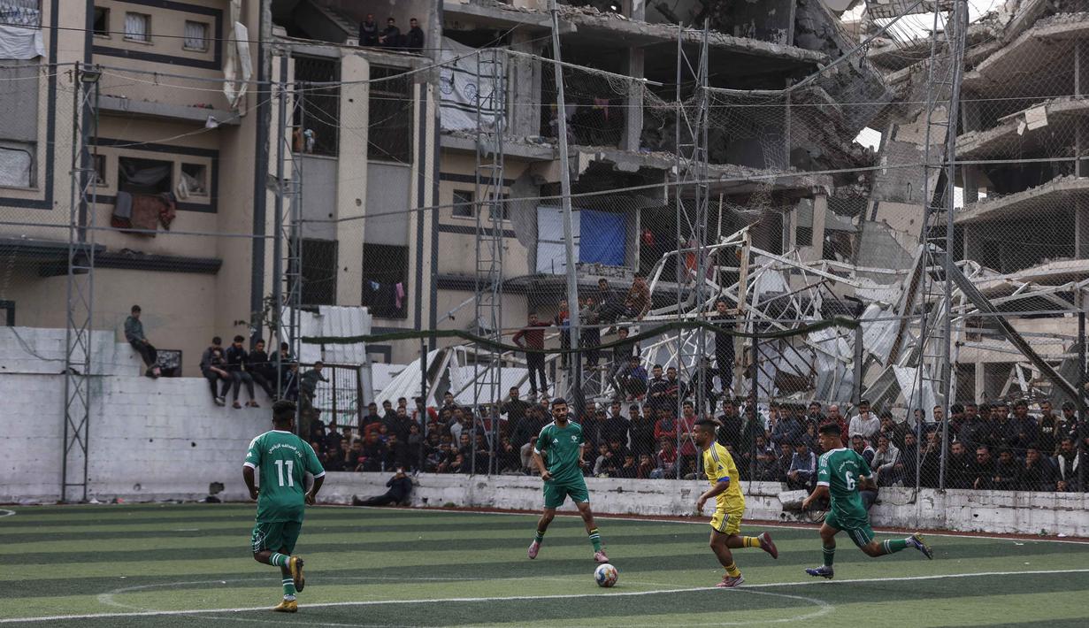 Setiap teriakan dari pinggir lapangan menjadi pengingat, di balik getirnya kehidupan di Gaza, ada jiwa-jiwa yang menolak menyerah. Tampak dalam foto, warga Palestina menyaksikan pertandingan sepak bola lima lawan lima di tengah reruntuhan bangunan yang hancur di Kota Gaza pada Senin 9 Februari 2026. (Omar AL-QATTAA/AFP)