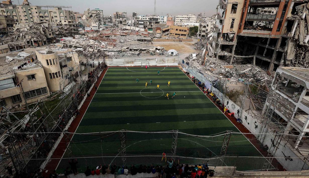 Sebuah pertandingan sepak bola lima lawan lima menarik perhatian warga Palestina di tengah kekerasan yang terus berlanjut di wilayah Gaza. Tampak dalam foto, warga Palestina menyaksikan pertandingan sepak bola lima lawan lima di tengah reruntuhan bangunan yang hancur di Kota Gaza pada Senin 9 Februari 2026. (Omar AL-QATTAA/AFP)