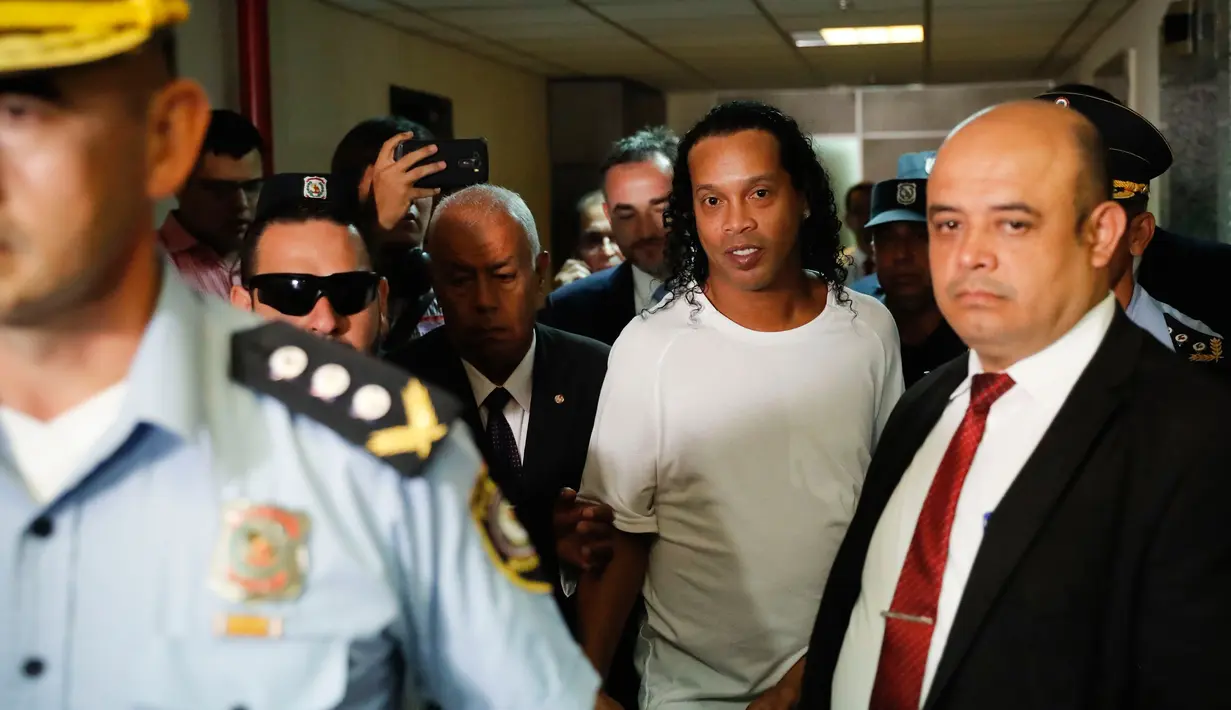 Mantan pemain timnas Brasil, Ronaldinho dikawal oleh polisi meninggalkan kantor pengadilan di Asuncion, ibu kota Paraguay, Jumat (6/3/2020). Ronaldinho bersama saudara laki-lakinya berurusan dengan pihak berwenang Paraguay karena menggunakan paspor palsu untuk memasuki negara itu. (AP/Jorge Saenz)