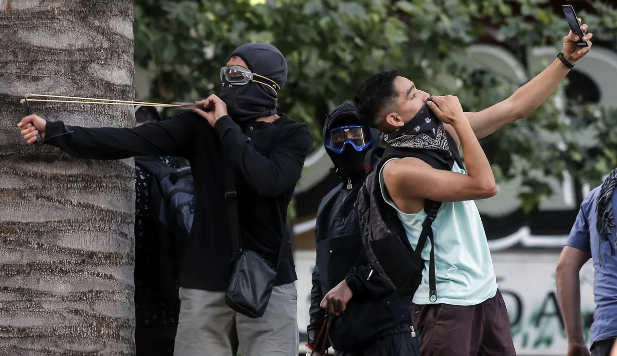 Demonstran berselfie ketika yang lain menggunakan ketapel menyerang polisi selama bentrok memprotes kebijakan ekonomi pemerintah di Santiago (6/11/2019). Presiden Chile mengatakan "tidak menyembunyikan apa-apa" mengenai tuduhan bahwa polisi membunuh, menyiksa warga sipil. (AFP Photo/Javier Torres)