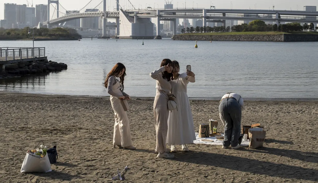 Wanita muda mengambil foto selfie pada hari yang cerah di tepi teluk di daerah Odaiba Tokyo (12/4/2022). Odaiba yang dapat diakses dari Jembatan Pelangi atau kereta Yurikamome yang futuristik adalah pusat hiburan teknologi canggih di sebuah pulau buatan di Teluk Tokyo. (AFP/Charly Triballeau)