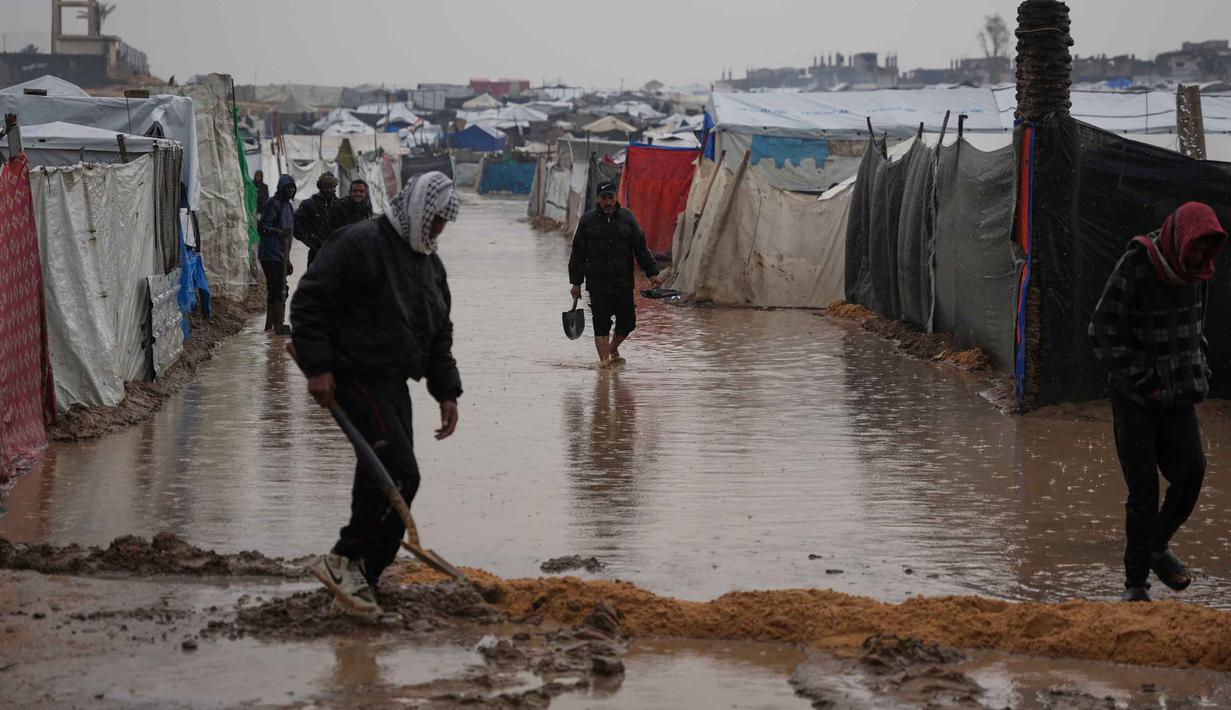 Puluhan hingga ratusan tenda pengungsi Palestina di wilayah tersebut terendam banjir. Tampak dalam foto, warga Palestina berjalan melewati kamp tenda yang terendam banjir setelah hujan lebat di Deir al-Balah, Jalur Gaza, Kamis 11 Desember 2025. (AP Photo/Abdel Kareem Hana)