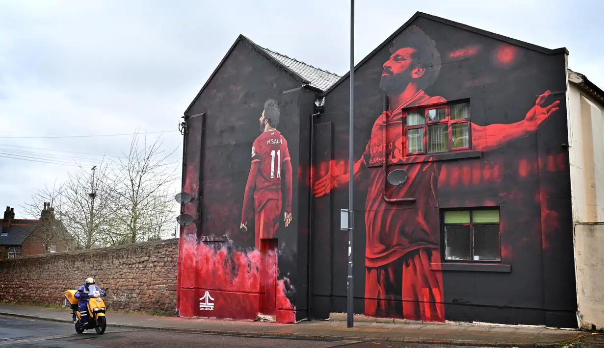 Pengendara skuter melewati mural seniman John Culshaw yang menggambarkan pesepak bola Mesir Mohamed Salah di fasad bangunan dekat Stadion Anfield, Liverpool, Rabu (30/3/2022). Salah tidak diragukan lagi, adalah salah satu pemain terhebat yang pernah mengenakan seragam Liverpool. (Paul ELLIS / AFP)