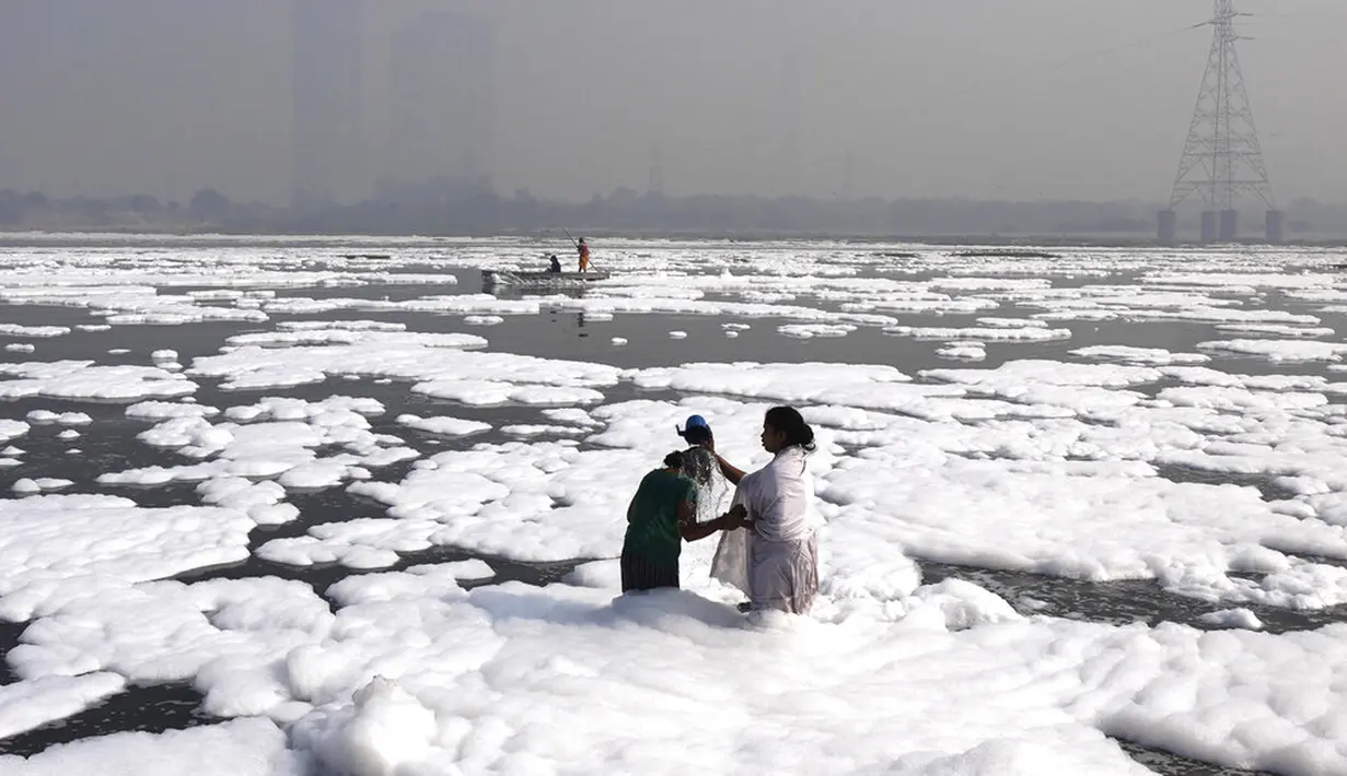 Seorang wanita memandikan putrinya di Sungai Yamuna yang tertutup busa kimia karena polusi industri dan domestik saat cakrawala diselimuti kabut asap beracun, New Delhi, India, 17 November 2021. India tutup sementara lima pembangkit listrik bertenaga batu bara karena polusi. (AP Photo/Manish Swarup)