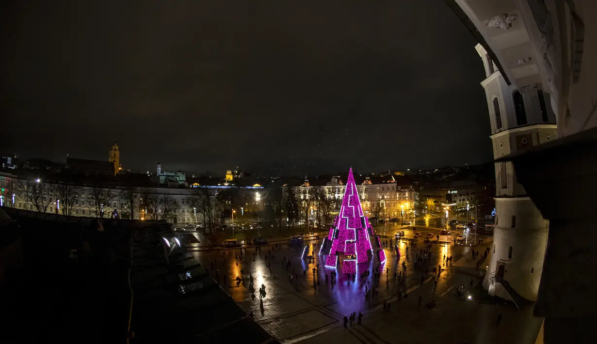 Orang-orang berjalan dekat pohon Natal Nasional selama upacara penyalaan di Lapangan Katedral yang sepi di Vilnius pada 28 November 2020. Pohon natal raksasa yang dihiasi berbagai ornamen lampu hias tersebut membuat kawasan di ibu kota Lithuania menjadi terang . (AP Photo/Mindaugas Kulbis)