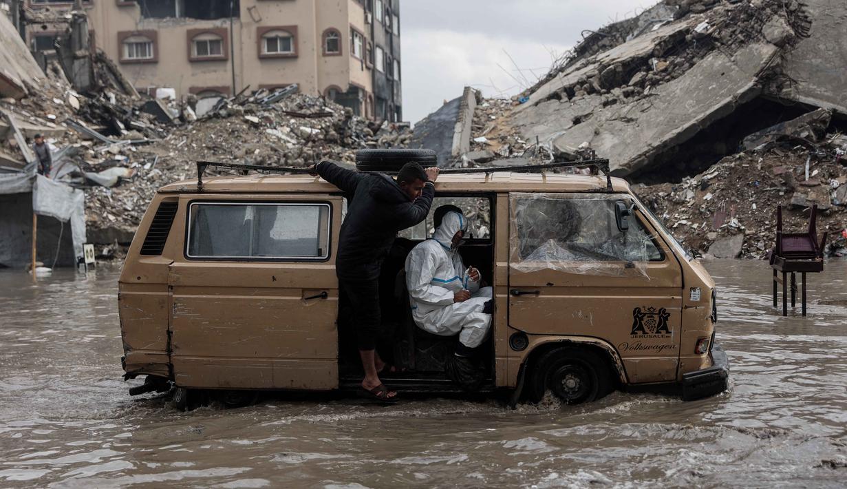 Sebagaimana diketahui, sebagian besar Kota Gaza hancur menjadi puing-puing dihantam serangan udara Israel sejak 7 Oktober 2023. Tampak dalam foto, orang-orang menaiki kendaraan yang dikendarai menerobos banjir di kamp darurat yang menampung warga Palestina yang terlantar setelah hujan deras di Kota Gaza pada Selasa 25 November 2025. (Omar AL-QATTAA/AFP)