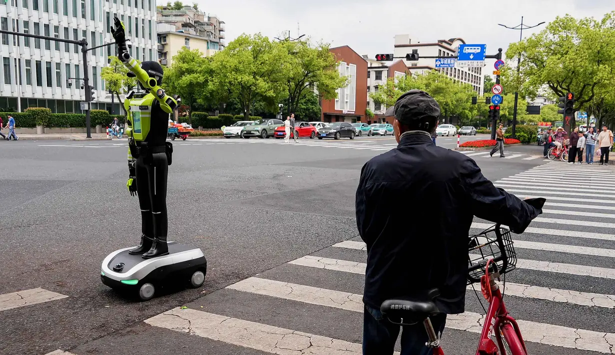 Robot-robot humanoid tersebut diterjunkan untuk membantu kepolisian mengatur arus lalu lintas dan memandu pejalan kaki, khususnya selama libur nasional Hari Buruh yang dimulai sejak 1 Mei 2026. Tampak dalam foto, seorang pesepeda melihat robot polisi humanoid di Hangzhou, provinsi Zhejiang Timur, Tiongkok pada Minggu 3 Mei 2026. (Agatha CANTRILL/AFP)