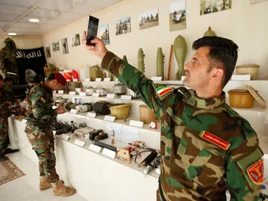 Seorang militer Kurdi Peshmerga berswafoto di sebelah bahan peledak dan barang-barang yang digunakan oleh militan Negara Islam (ISIS) di sebuah museum di Erbil, Irak, 12 Mei 2019. 'Museum ISIS' tersebut dibuka oleh militer Peshmerga. (REUTERS/Azad Lashkari)