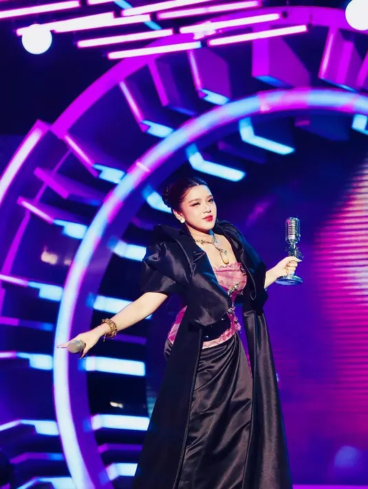 Tiara Andini tampil di panggung mengenakan outfit edgy berwarna hitam. Outfit ini memiliki atasan bernuansa keunguan. Tiara juga memadukan penampilannya dengan makeup bold, rambut ditata sleek dengan high-bun, dan aksesori emas yang serasi. Foto: Instagram.