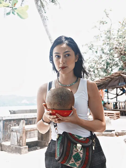 Berlibur di Ambon, Clara tidak melewatkan kesempata untuk mencoba kelapa muda segar di pantai. Menggunakan tank top putih, dengan celana hitam, penampilannya semakin lengkap dengan tas sandang bermotif khas nusantara. (Liputan6.com/IG/clarabernadeth)
