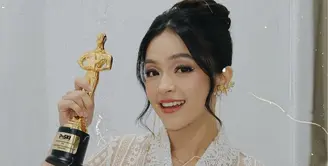 Wajahnya dipoles dengan blush tipis coral senada dengan warna lipstik glossynya. Riasan mata dibuat soft dengan shimmer dan garis putih di kelopak mata dan sedikit eyeliner wings. Rambutnya ditata sanggul modern tanpa hiasan dengan poni membingkai wajah. [elsaajapasal]