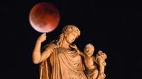 Foto ini menampilkan bulan purnama yang juga dikenal sebagai “Blood Moon” selama gerhana bulan, dengan patung dewi Yunani kuno Irene yang memegang seorang anak di latar depan, di pusat Athena, Yunani pada 7 September 2025. (Angelos TZORTZINIS/AFP)