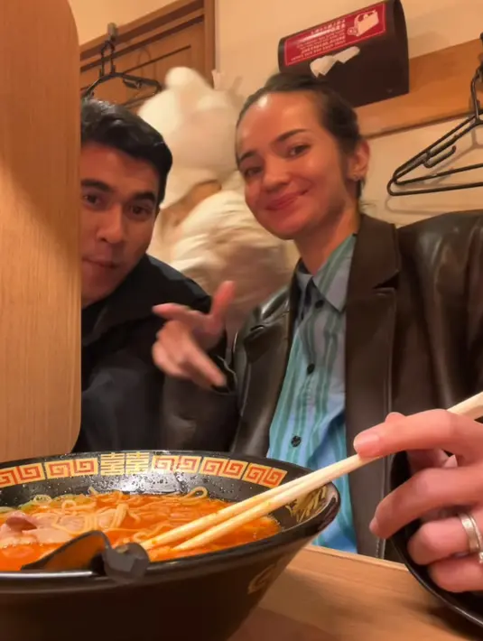 Enzy mengenakan jaket kulit coklat dipadukan inner kemeja warna biru dan hijau saat menyantap ramen bersama suaminya. [@enzystoria]