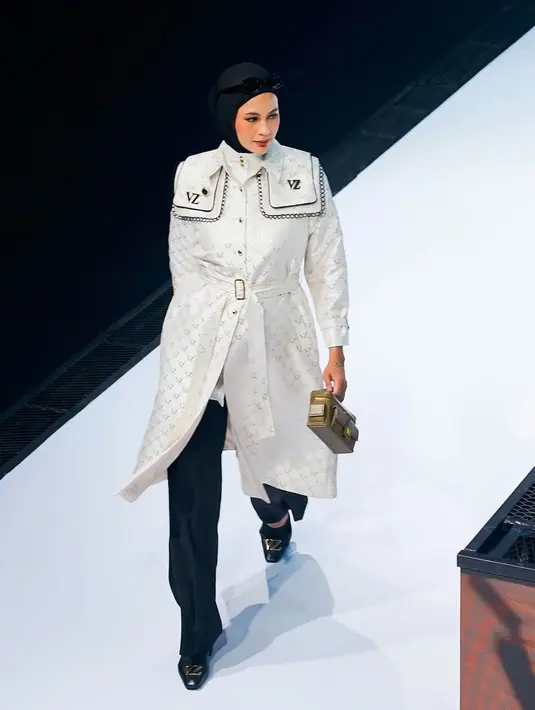 Paula mengenakan trench coat putih dengan monogram khas Vivi Zubedi yang dipadukan dengan celana hitam panjang.  [@vivizubedi].