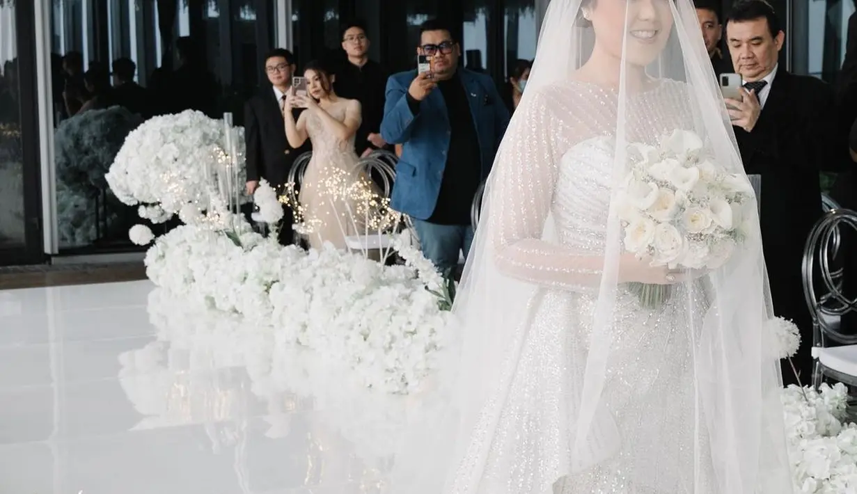 Ia tampil cantik dengan gaun putih saat memasuki altar. Gaun ini menggabungkan strapless dress dan lengan panjang transparan yang sopan   [Foto: Instagram/@sekisah.photo/@sekisah.sangjit].