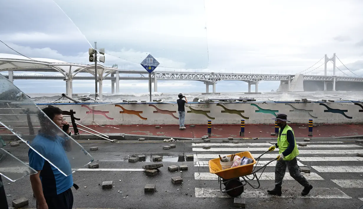 Seorang pekerja (kanan) terlihat di balik jendela kaca yang pecah saat ia mendorong gerobak dorong melewati batu paving di jalan, setelah Topan Hinnamnor melewati Busan, Korea Selatan (6/9/2022). (AFP/Anthony Wallace)