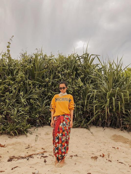 Indah berpose di sebuah pantai menggunakan sweater kuning dan batik berwarna merah bercorak. Ditambah kacamata hitam, dia tampak percaya diri dengan background semak dan langit kelabu. (Liputan6.com/IG/indahpermatas)