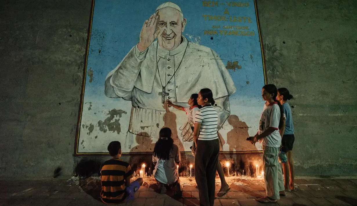 Paus Fransiskus lahir dengan nama Jorge Mario Bergoglio di ibu kota Argentina, Buenos Aires, pada tahun 1936. Tampak dalam foto, umat Katolik berduka atas kematian Paus Fransiskus dengan menyalakan lilin di depan muralnya di dekat Esplanade Tasitolu, tempat Paus mengadakan Misa pada September tahun lalu, di Dili, Timor Leste, pada 22 April 2025. (YASUYOSHI CHIBA/AFP)