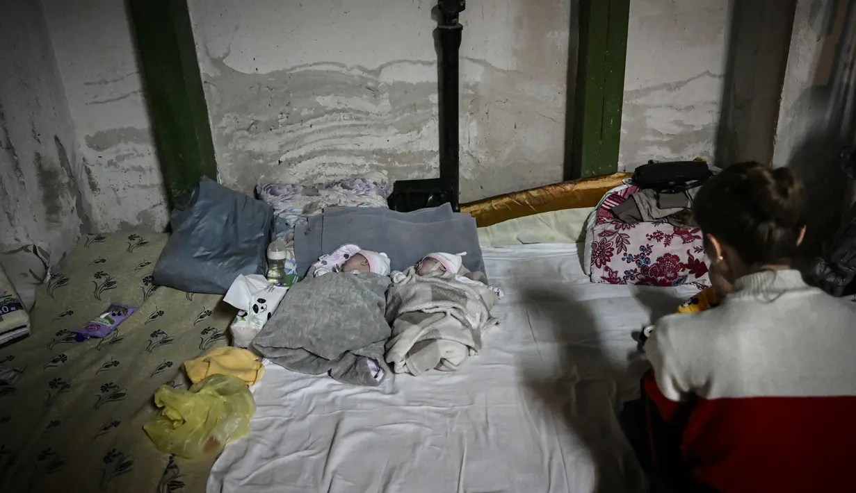 Seorang perempuan duduk dengan dua bayi yang dirawat di rumah sakit pediatri setelah dipindahkan ke ruang bawah tanah yang digunakan sebagai shelter bom di Kiev, Senin (28/2/2022). Tentara Rusia mengatakan, warga sipil Ukraina dapat meninggalkan ibu kota Kiev dengan bebas. (Aris Messinis/AFP)