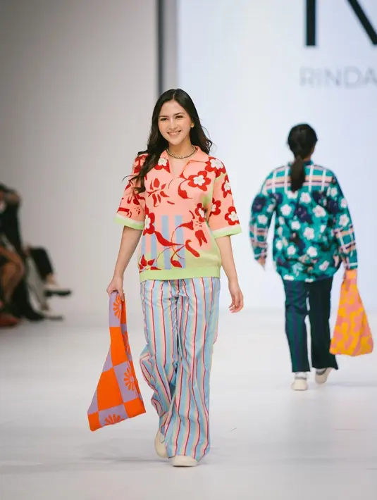 Jessica Mila melenggang di runway mengenakan atasan rajut berkerah berwarna pink bermotif bunga yang cantik. Ia padukan dengan celana biru bermotif garis dan menenteng tas rajut oranye. Foto: Instagram.