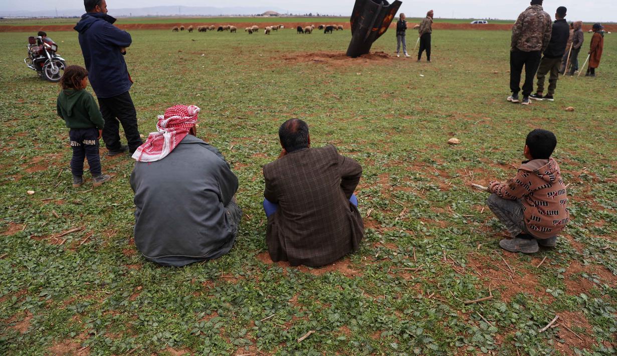 Keberadaan proyektil tersebut menarik perhatian penduduk lokal, termasuk anak-anak dan penggembala muda, untuk mendekati dan melihat dari dekat. Tampak dalam foto, warga setempat melihat proyektil rudal Iran yang belum meledak di lapangan terbuka di pinggiran Qamishli, Suriah timur, Rabu 4 Maret 2026. (AP Photo/Baderkhan Ahmad)