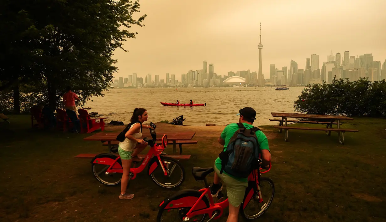 Warga berhenti sejenak saat bersepeda di Kepulauan Toronto dengan latar cakrawala pusat kota yang diselimuti asap akibat kebakaran hutan di Ontario Utara, yang menyebabkan kualitas udara memburuk, di Toronto, Ontario, Kanada, Kamis, 15 Juli 2026. (AFP/ Cole Burston)