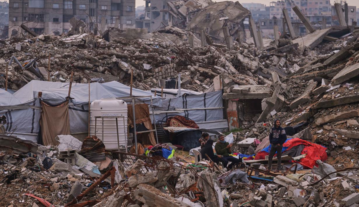Warga Palestina berdiri di samping tenda yang didirikan di atas reruntuhan bangunan yang hancur akibat serangan udara dan darat Israel di lingkungan Sheikh Radwan, Kota Gaza, Selasa, 30 Desember 2025. Setelah lebih dari dua tahun agresi militer Israel yang menghancurkan wilayah tersebut, kini cuaca dingin ekstrem dan hujan lebat menjadi tantangan besar bagi pengungsi di Jalur Gaza. (AP Photo/Abdel Kareem Hana)